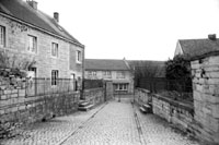 Allée principale pavée conduisant à l’église et sa barrière (1975). A gauche : le presbytère encore entouré de grillages ; à droite : accès au jardin du presbytère.